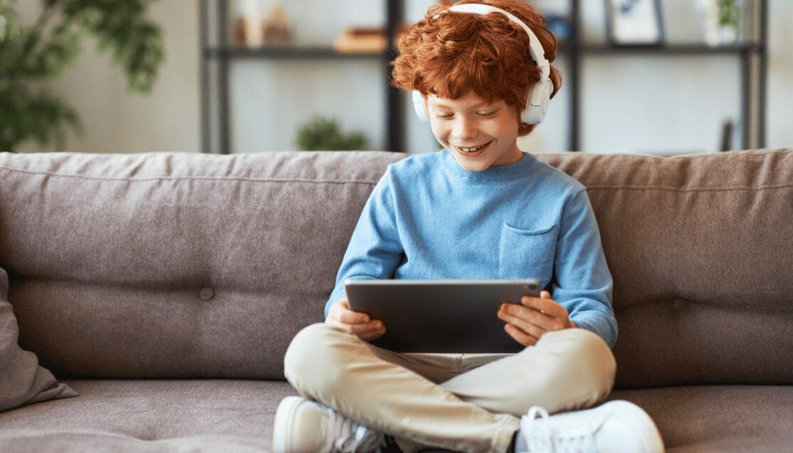 A boy with red hair sits on a couch, focused on a tablet in his hands, surrounded by a cozy living room setting.