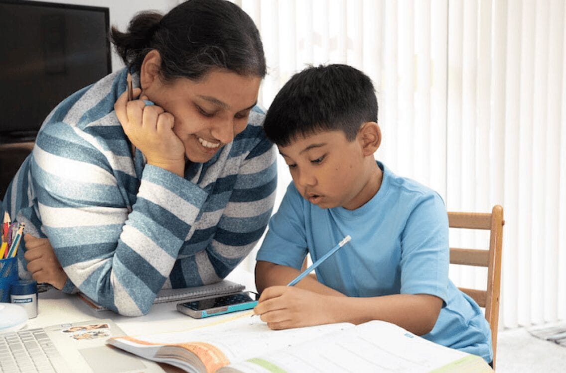 A woman and a boy collaborate on homework, focused on their studies at a table filled with books and papers.