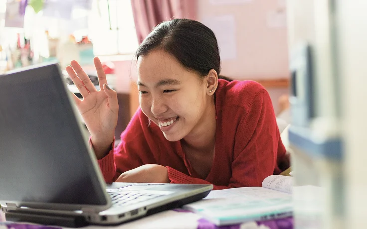Una mujer sonriente con un suéter rojo saluda la pantalla de una computadora portátil, transmitiendo una interacción alegre. Está acostada en una cama con un libro cerca en una habitación acogedora.