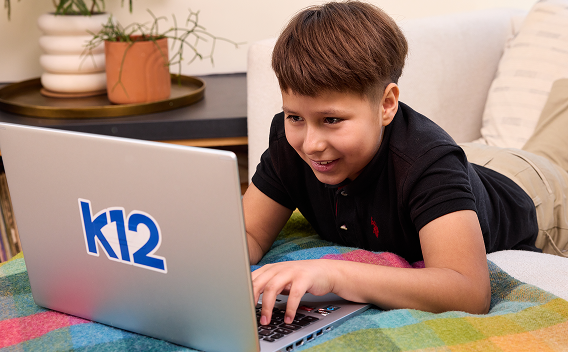 A young boy, smiling and focused, is lying on a couch using a laptop with a k12 logo. He appears engaged with his colorful surroundings.