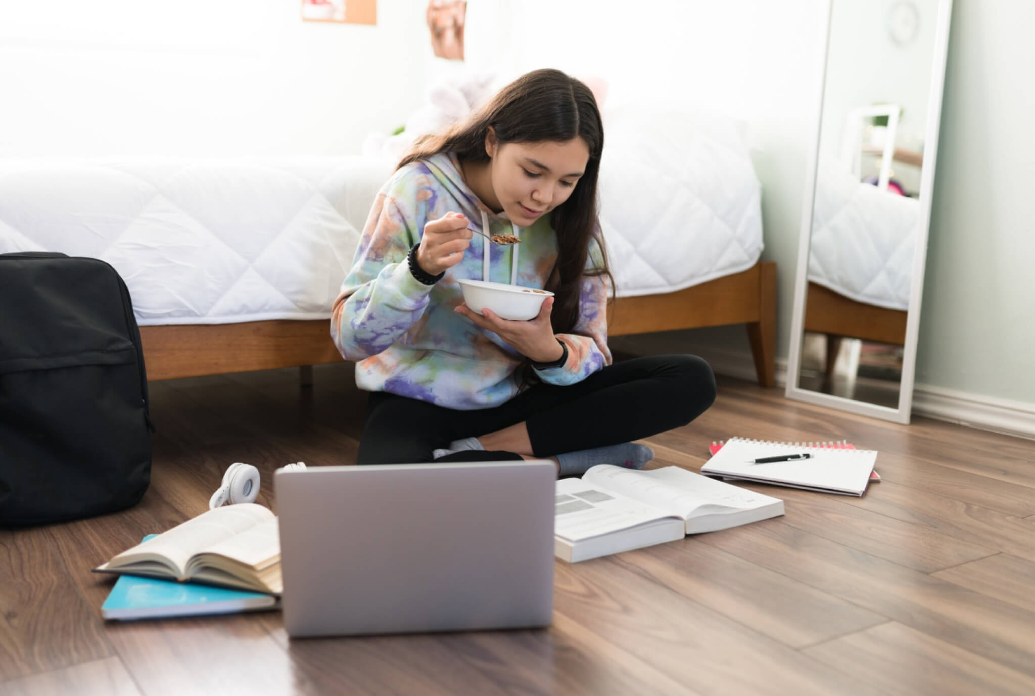 A young girl sits cross-legged on a wooden floor, eating cereal while studying. Open books, a notebook, and a laptop surround her, with a bed and mirror in the background.