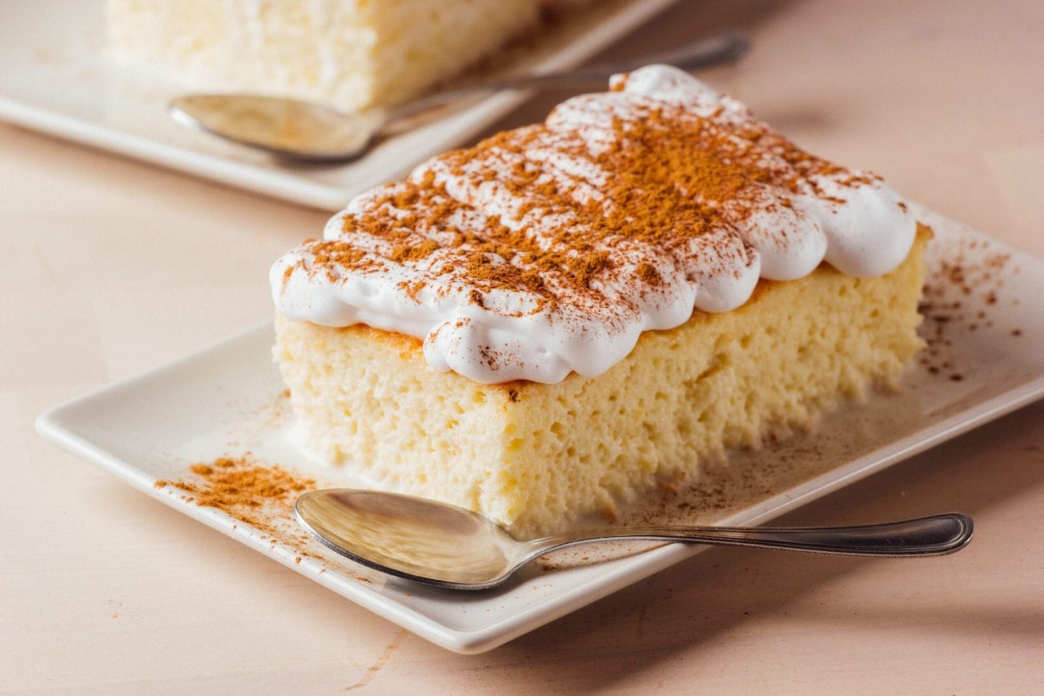 Rebanada de pastel de tres leches cubierto con crema batida y canela en un plato blanco con una cuchara. El fondo claro agrega una sensación acogedora y acogedora.