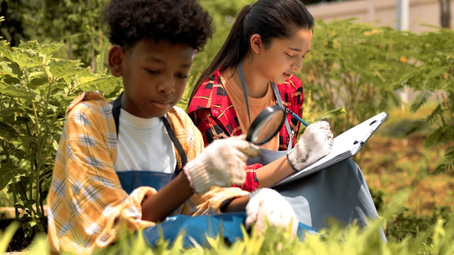 Two children in a garden; one observes plants with a magnifying glass, the other records notes on a clipboard. They wear gloves and aprons, focused and engaged.