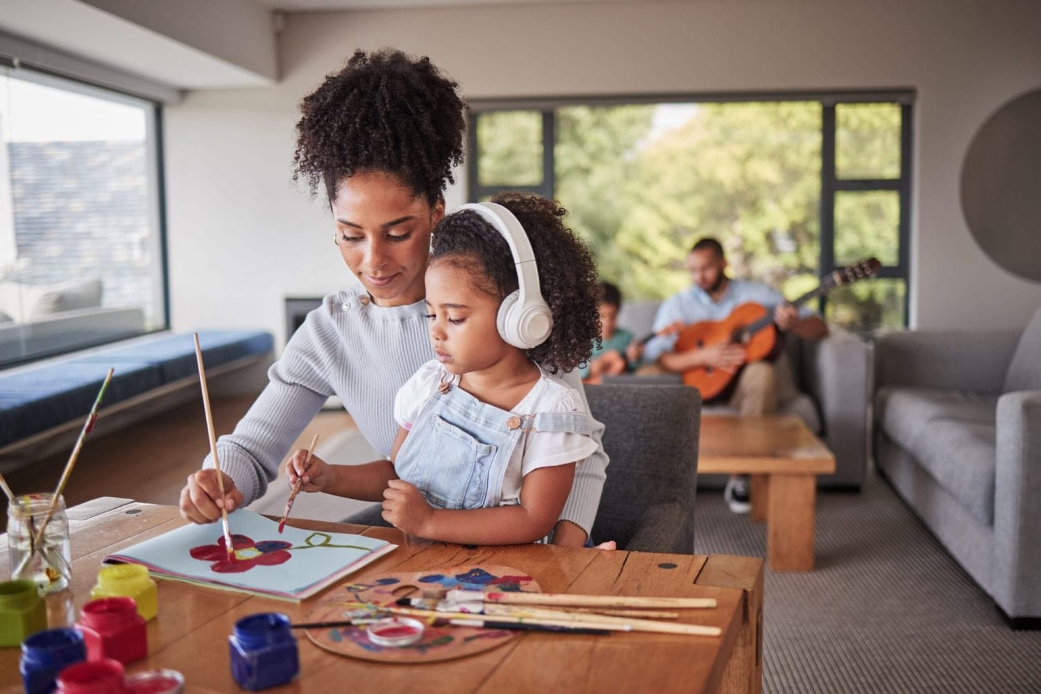 Mother and daughter paint together at a wooden table, the girl wearing headphones. A man plays guitar in a cozy, sunlit living room. Family bonding.