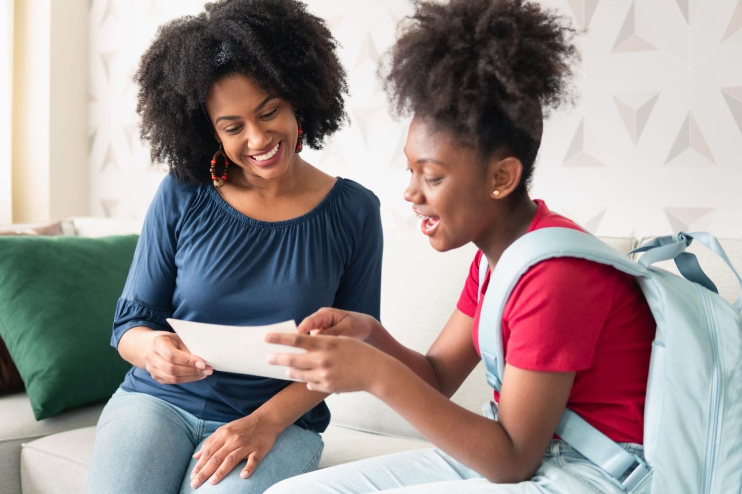 A woman in a blue top and a young girl with a backpack share a happy moment on a couch, smiling while looking at a paper, conveying joy and connection.