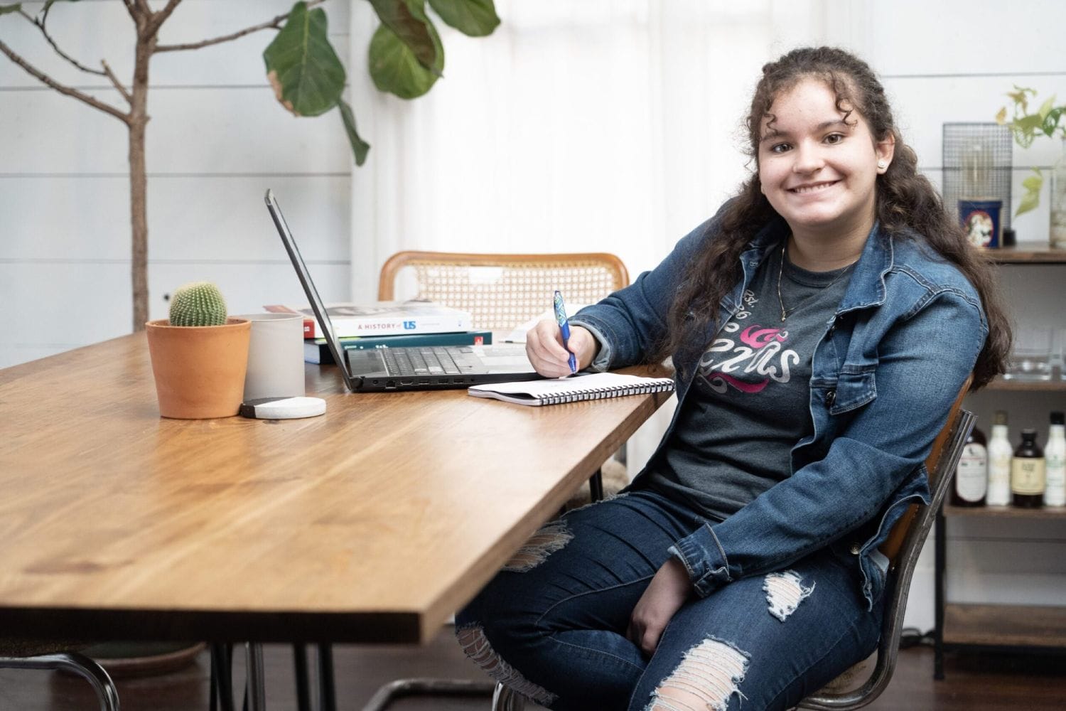 Mujer joven sonriendo, sentada en una mesa de madera con una computadora portátil, libros y un cuaderno. Ella usa un atuendo de mezclilla, transmitiendo un ambiente de estudio relajado.