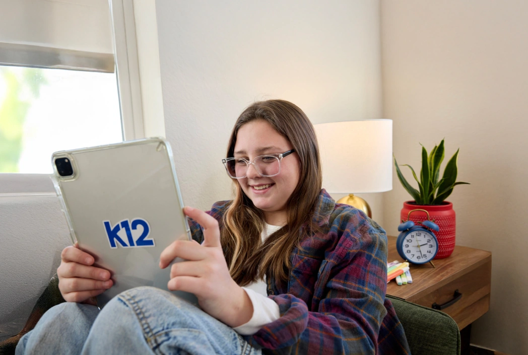 A girl with long hair and glasses smiles while using a tablet labeled "k12" in a cozy room with a lamp, plant, and clock on a wooden table.