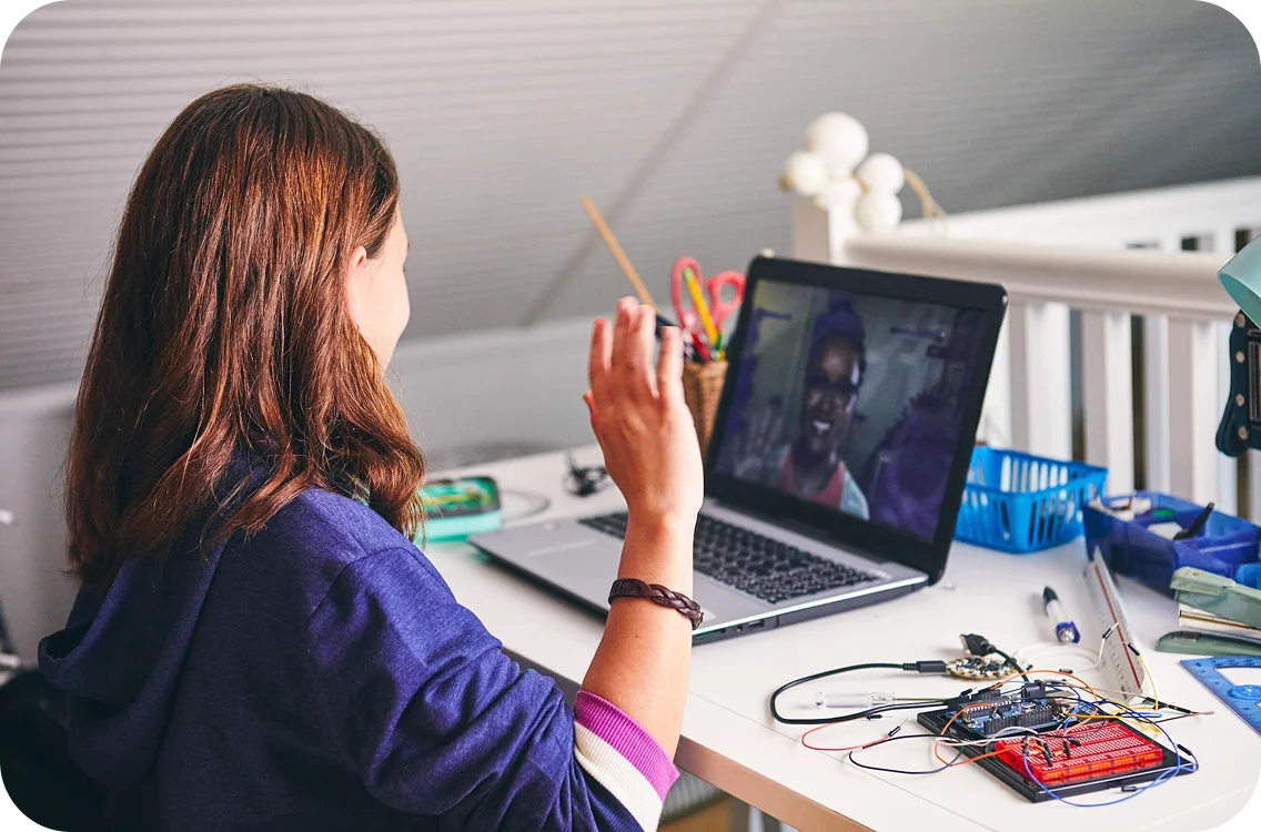 A person with brown hair waves at a laptop during a video call. Electronics components are spread on the desk. The setting is casual and tech-focused.