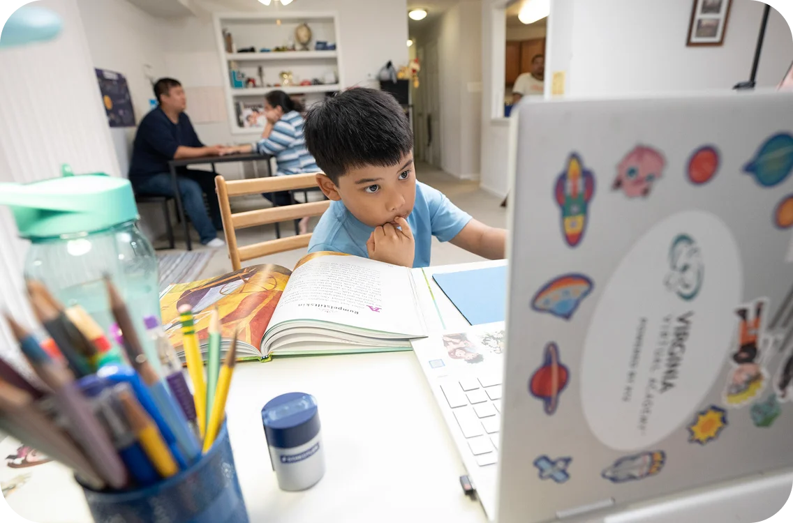 A young boy focuses intently on a laptop with colorful stickers, surrounded by open books and pencils. Two adults converse at a table in the background.
