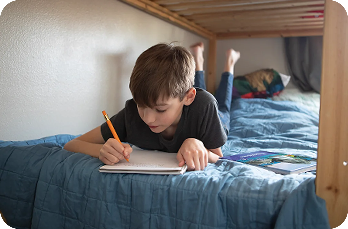 A young boy lies on his bed, concentrating while writing in a notebook. He holds a pencil and is surrounded by books, creating a focused and studious atmosphere.