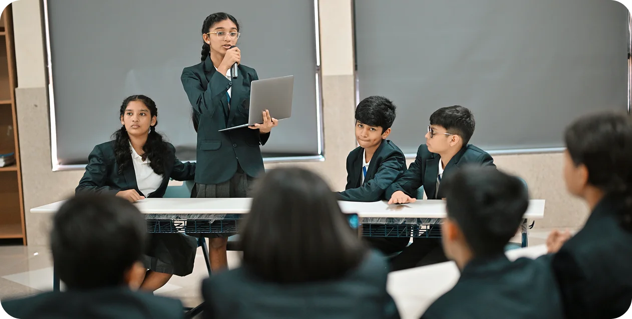 A group of students in uniforms sit at tables while a standing student speaks confidently, holding a laptop. The mood is focused and collaborative.
