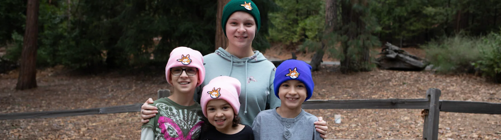 A group of four smiling children stand outdoors in a forest. They wear colorful beanies featuring animal designs. The mood is cheerful and relaxed.
