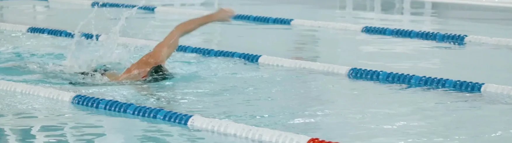 A swimmer performs the freestyle stroke in a clear swimming pool lane marked with blue and white lane dividers, creating splashes and conveying motion.