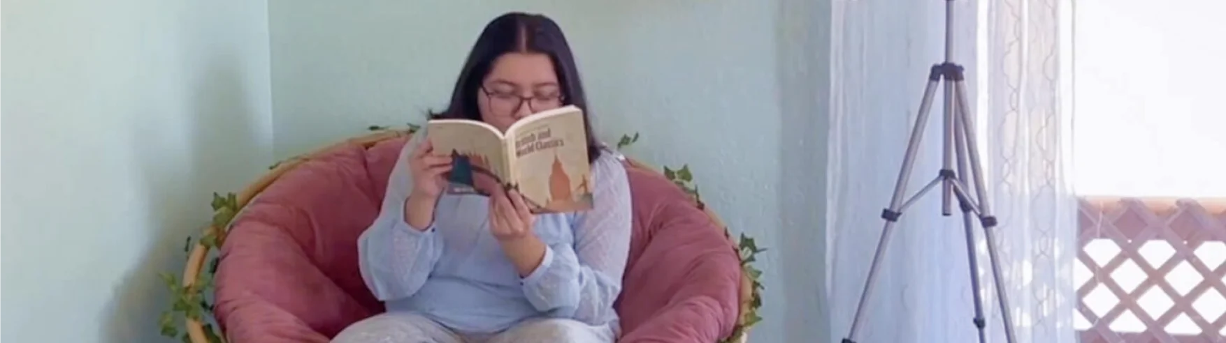 A person with long dark hair sits on a large, cushioned chair, reading a book titled "The Little Prince." They appear relaxed in a cozy, softly lit room.