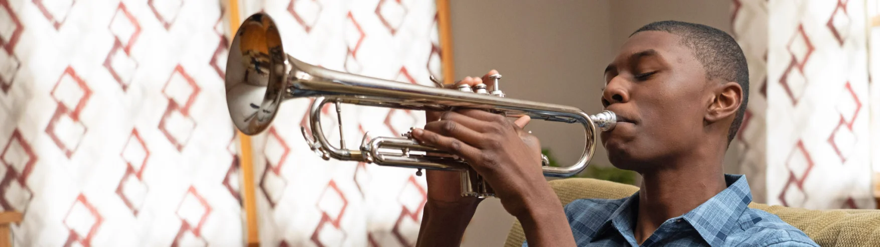 A person sits on a couch playing a silver trumpet, eyes closed in concentration. The background features curtains with a geometric pattern, creating a calm ambiance.