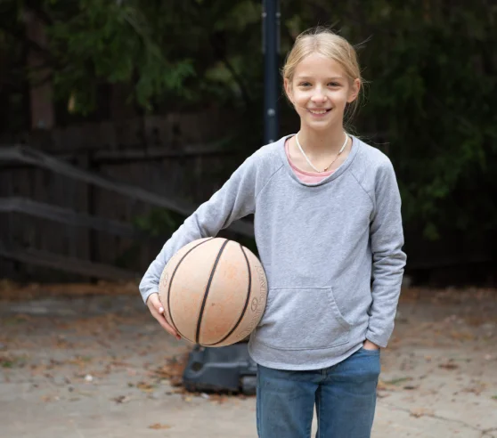 A young girl stands outdoors, holding a basketball under one arm. She wears a gray hoodie and jeans, smiling against a background of trees and a wooden fence.