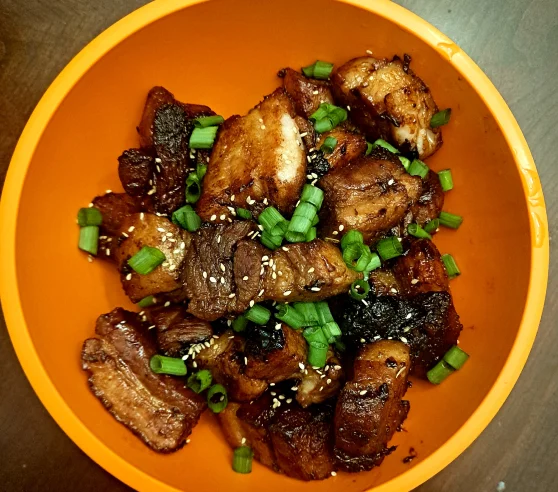 A vibrant orange bowl filled with succulent, crispy pork belly chunks garnished with sesame seeds and fresh green onion slices.