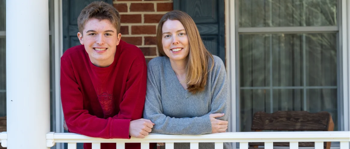 A young man in a red sweater and a woman in a gray sweater lean on a porch railing, smiling. The background features brick walls and windows, creating a cozy atmosphere.