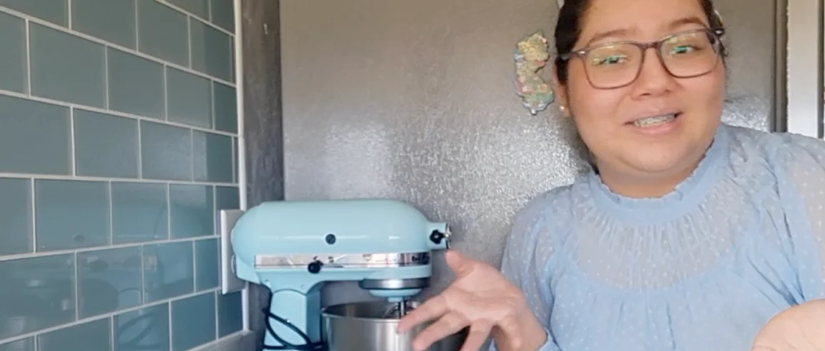 A woman with glasses and a blue blouse gestures while smiling in a kitchen. Beside her is a light blue stand mixer on a countertop with blue tile.