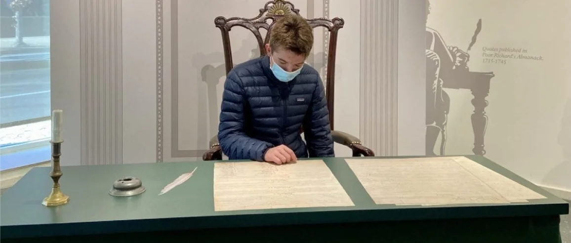 A person wearing a mask sits at a green desk with historical documents, focused intently. A candle and quill nearby suggest a scholarly setting.