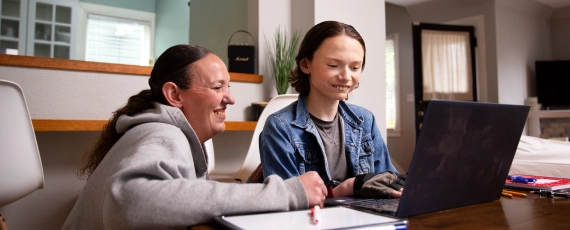 A cheerful woman and a young boy sit at a table, engaged with a laptop. The room is cozy and well-lit, conveying warmth and collaboration.