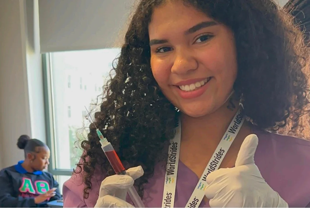 Young woman smiling confidently, wearing gloves and holding a syringe filled with red liquid. She's in a bright room, another person is visible in the background.