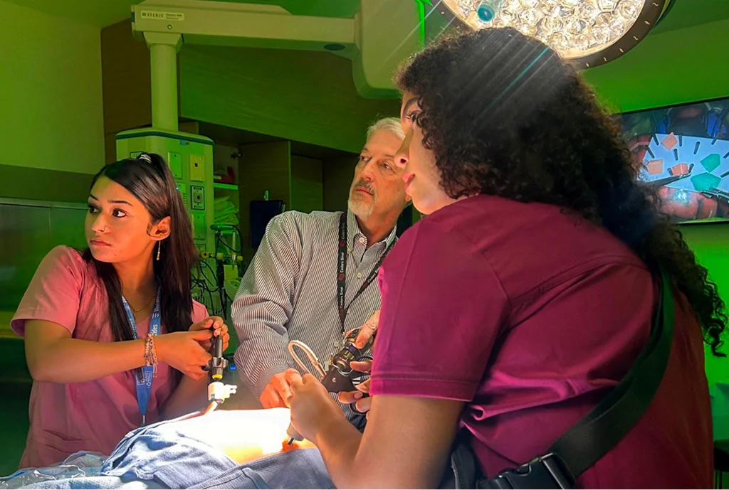 A group of three medical professionals in a brightly lit operating room, focused on a procedure. The scene conveys teamwork and concentration.