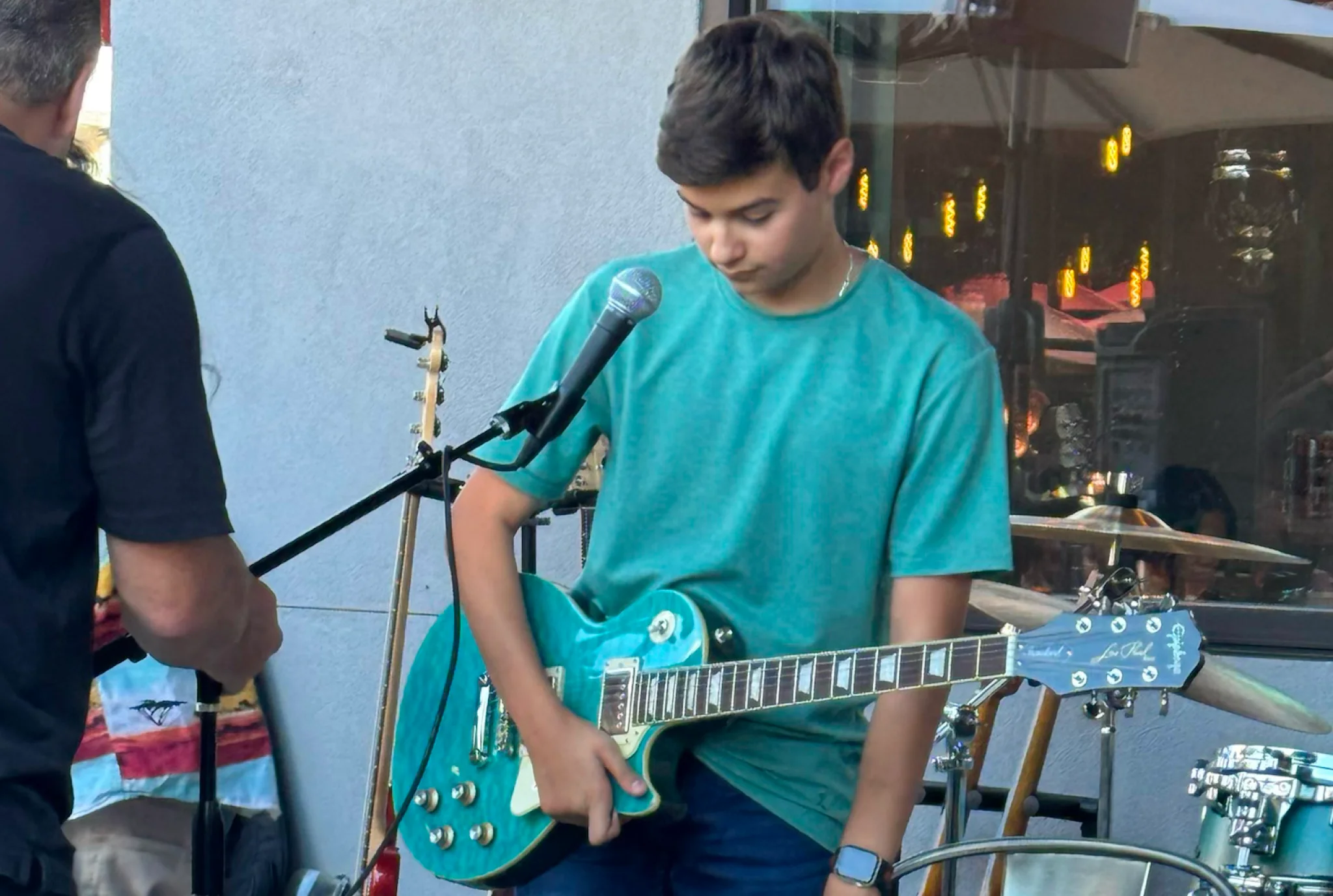 Niño con una camisa verde azulado afinando una guitarra eléctrica azul en el escenario. Un micrófono y batería están cerca. Ambiente casual y centrado.