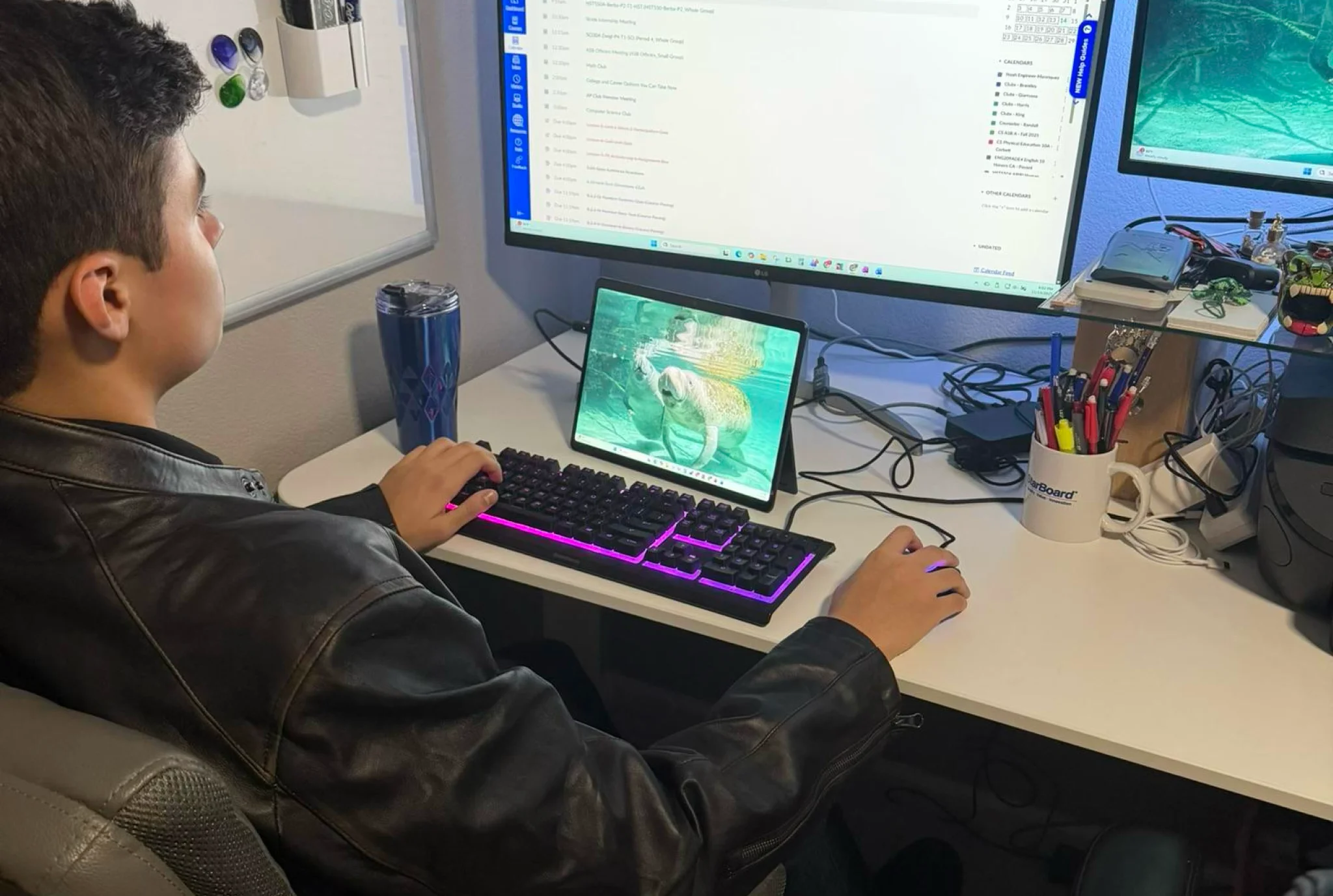 Una persona con una chaqueta de cuero se sienta en un escritorio, usando una computadora con una configuración de doble monitor y una tableta que muestra un pez. La escena es enfocada y productiva.