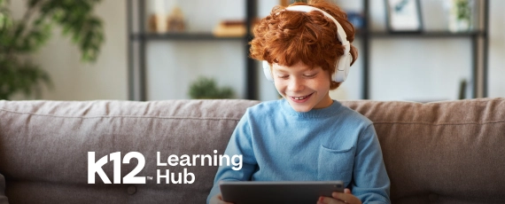 Young boy with curly red hair wearing headphones, smiling at a tablet. He sits on a couch in a cozy room. "K12 Learning Hub" text is visible.