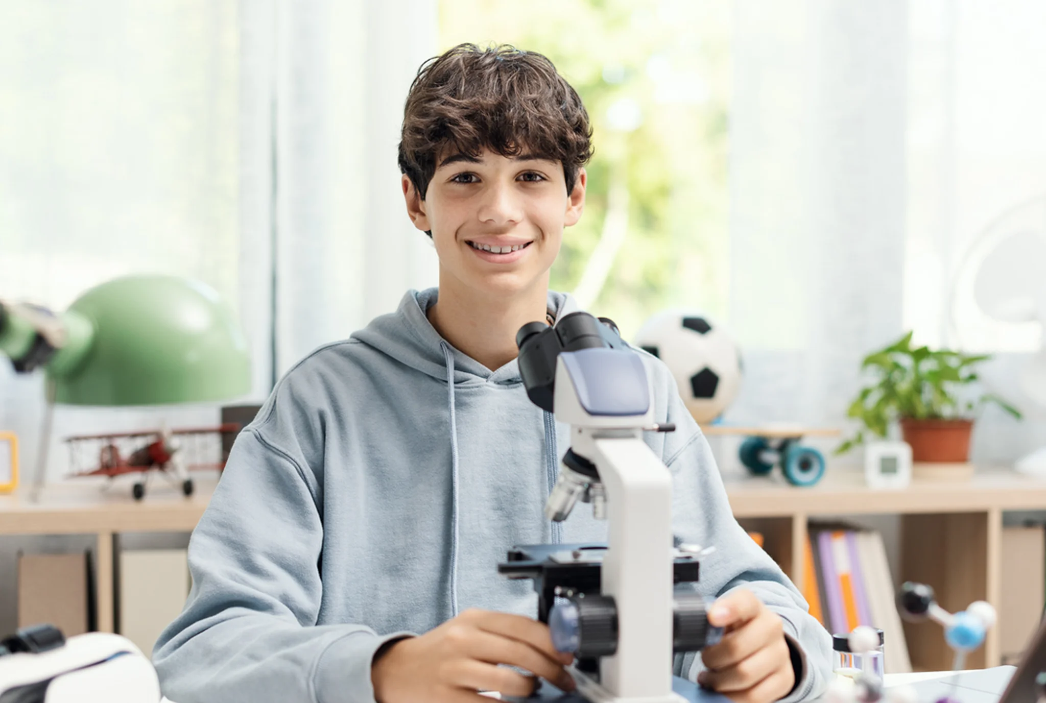 Un adolescente con cabello castaño rizado sonríe detrás de un microscopio en una habitación luminosa y acogedora. Los estantes con un balón de fútbol y una planta son visibles.