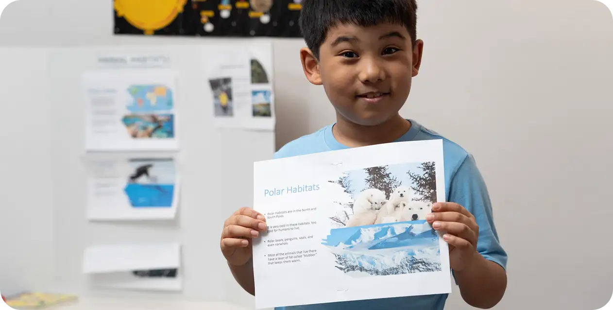Young boy holding a paper with "Polar Habitats" text and polar bear images. He's smiling, with educational posters in the background, conveying learning and curiosity.