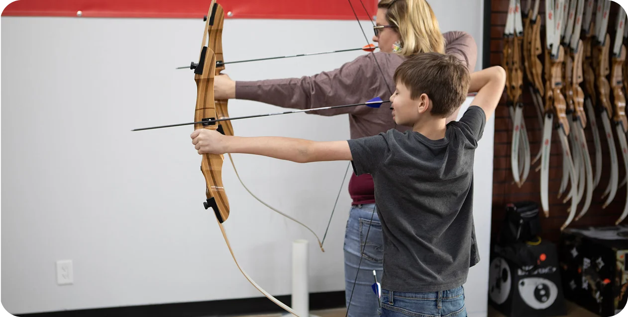 A boy and a woman practice archery indoors, aiming wooden bows at a target off-frame. The atmosphere is focused, with hanging bows in the background.