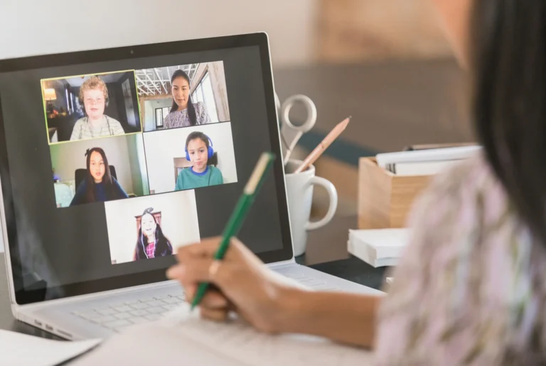 A person takes notes with a pencil while attending a video conference on a laptop. The screen shows five people in a virtual meeting.