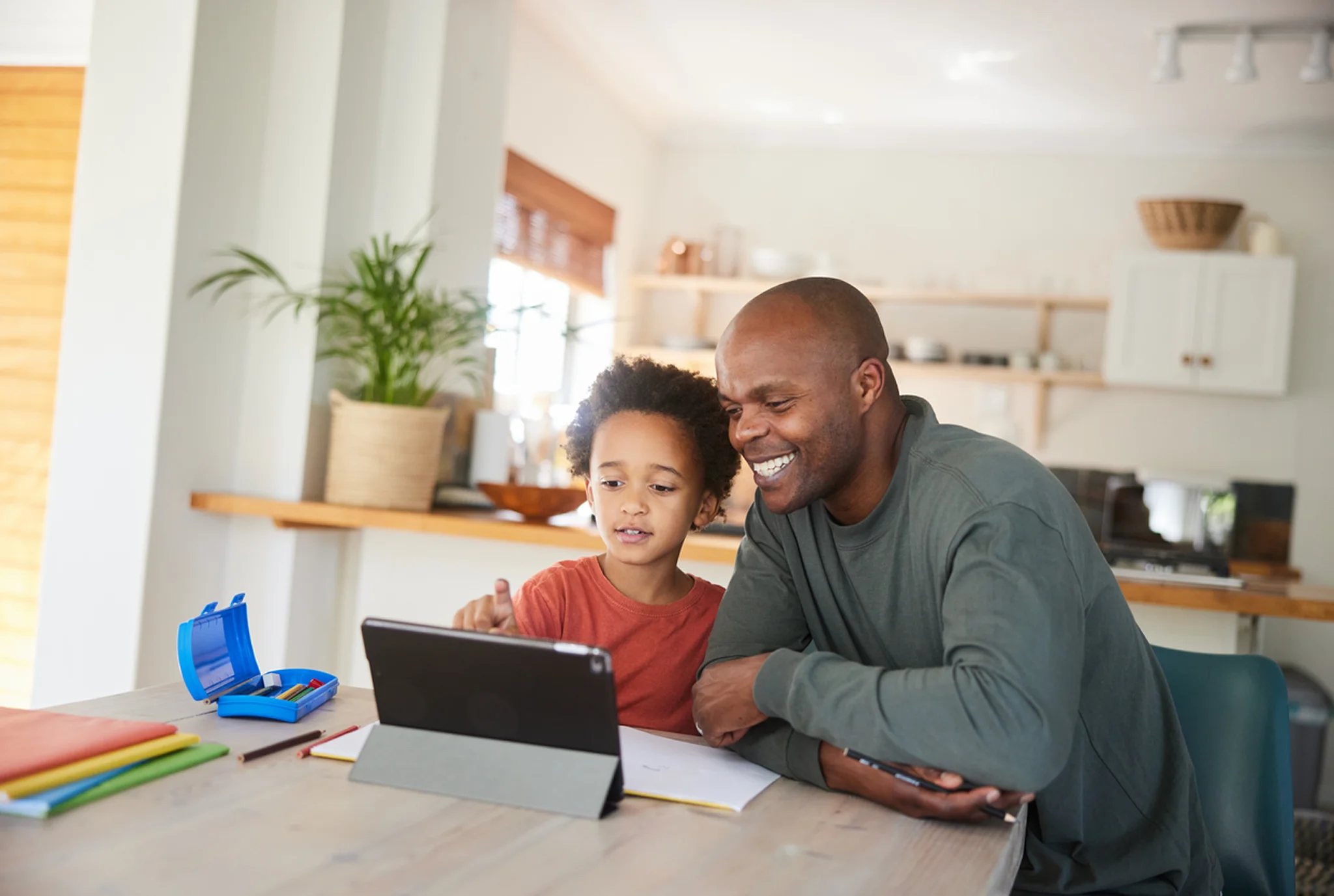 Padre e hijo se sientan en la mesa de la cocina, sonriendo mientras usan una tableta juntos. El entorno es cálido y acogedor, con una cocina moderna en el fondo.