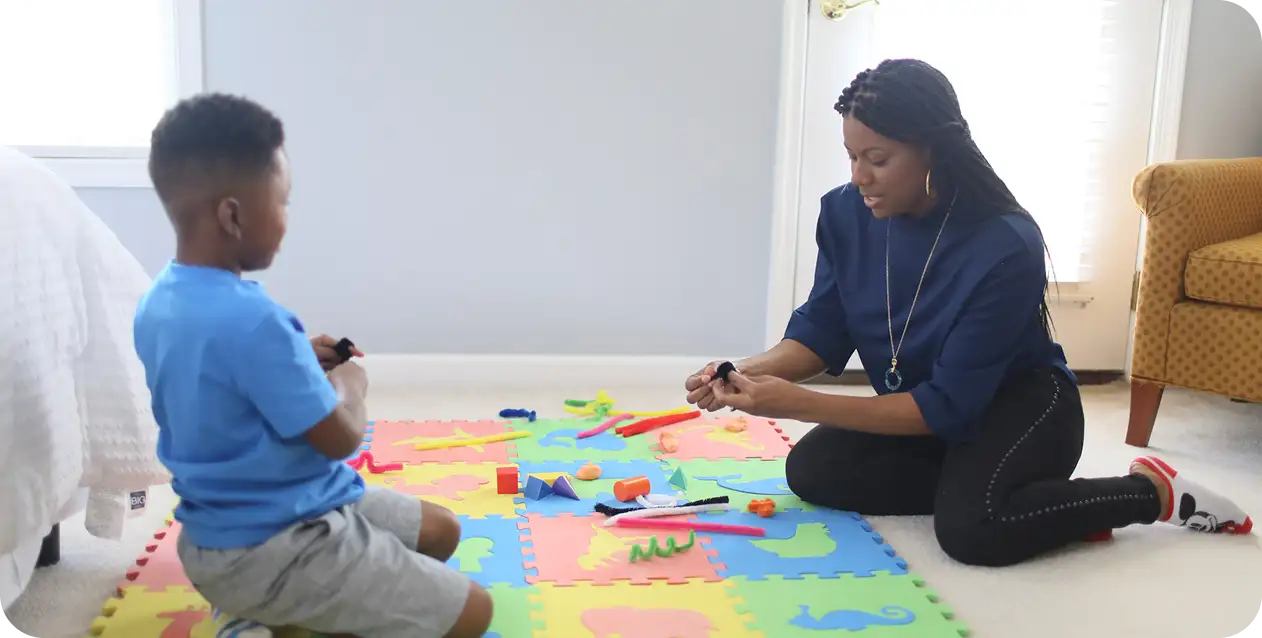 A woman and a child sit on colorful foam mats, playing with toy blocks and tools. The room is bright, creating a playful and warm atmosphere.