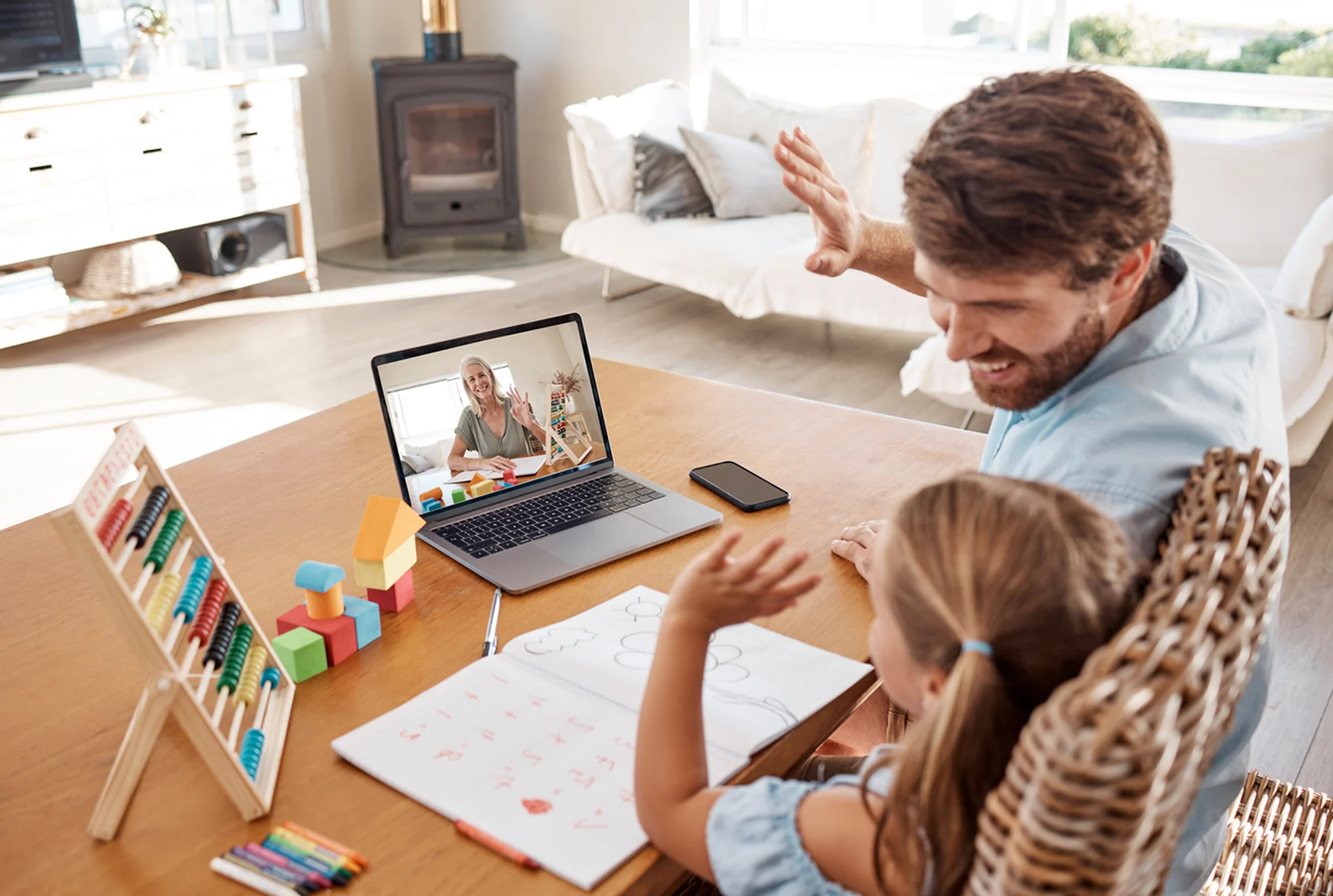 Father and daughter in a cozy living room wave at a laptop screen during an online lesson. A teacher smiles on the display. A warm, educational setting.