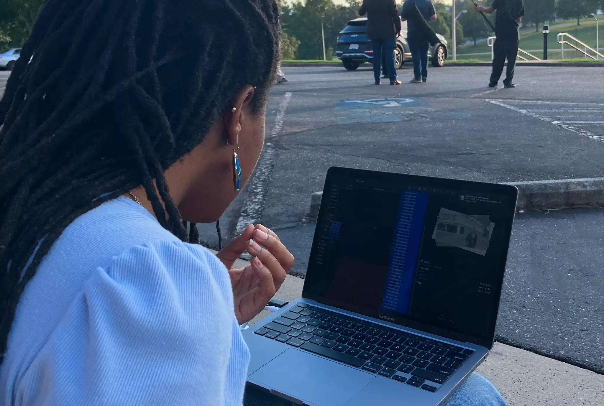 Una persona con cabello largo y aretes azules se sienta al aire libre usando una computadora portátil que muestra una imagen 3D. En el fondo, la gente se para cerca de un automóvil estacionado en una carretera.