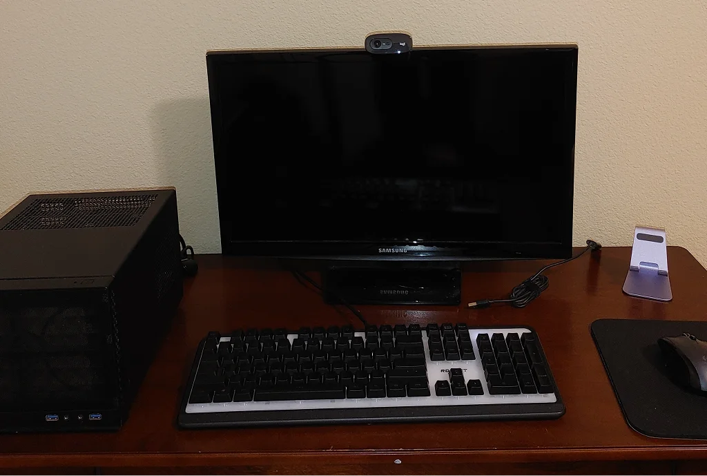 A computer setup on a wooden desk with a monitor, keyboard, and mouse. A small black computer case sits to the left, and a phone stand is on the right.