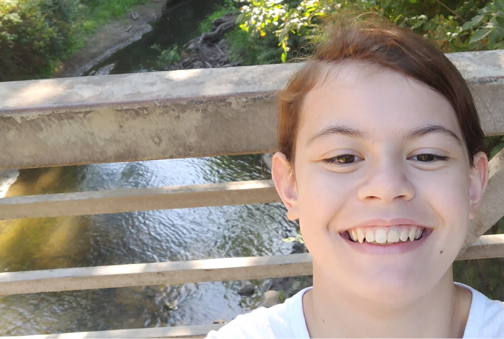 Smiling person taking a selfie on a bridge, with a calm river and lush greenery in the background. The mood is cheerful and relaxed.