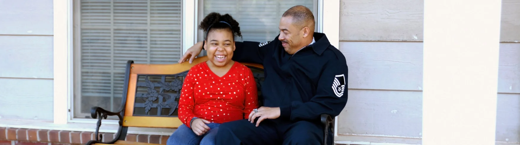 Father and daughter smiling on a porch bench. The father, in a dark uniform, lovingly wraps his arm around his daughter in a red polka dot shirt. Relaxed, joyful moment.
