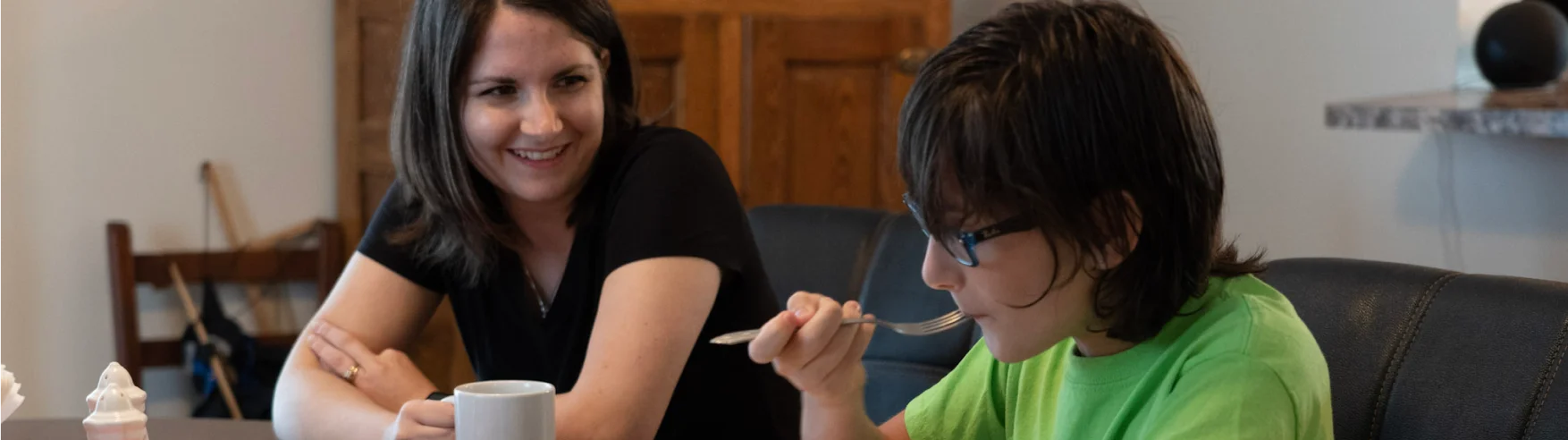 A woman smiles while watching a boy in glasses eat at a table. The boy wears a green shirt, focused on his food. The mood is warm and content.