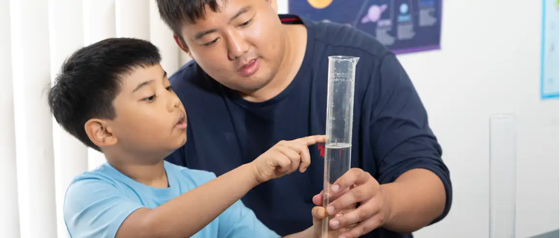 A young boy and a man focus intently on a graduated cylinder in a bright room, suggesting a science lesson. The scene conveys curiosity and learning.