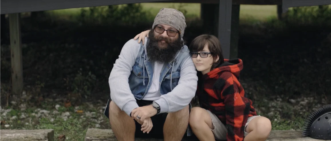 A bearded man and a child sit closely, smiling warmly. The man wears a beanie and glasses, while the child sports a red plaid jacket. The atmosphere is cozy.