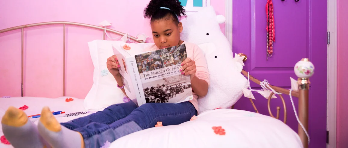 A girl relaxes on a bed with floral bedding in a pink room, reading a history book. A fluffy toy sits beside her, creating a cozy atmosphere.