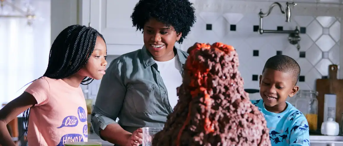 A woman and two children smile at a large erupting volcano model in a kitchen. The scene conveys excitement and curiosity in a joyful atmosphere.