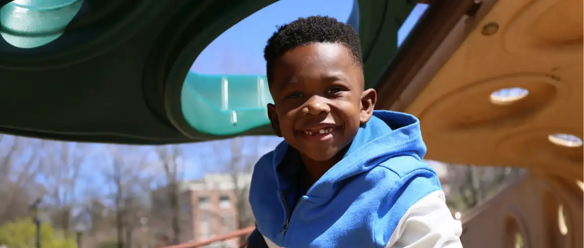 A young boy in a blue hoodie smiles brightly while playing at a playground. The sunny day and green play structure evoke a cheerful and energetic mood.