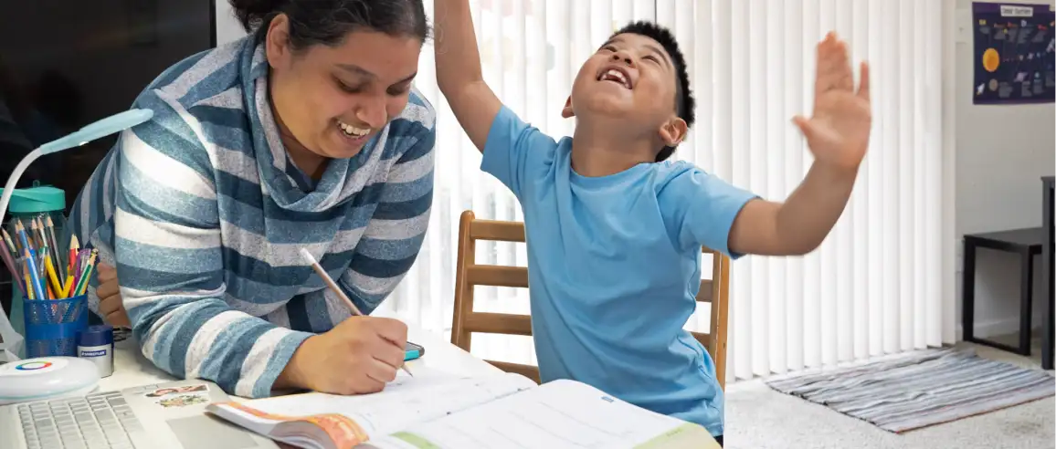 A woman and a young boy share a moment at a table. She's smiling while writing in a notebook, and the boy, in a blue shirt, raises his arms joyfully.