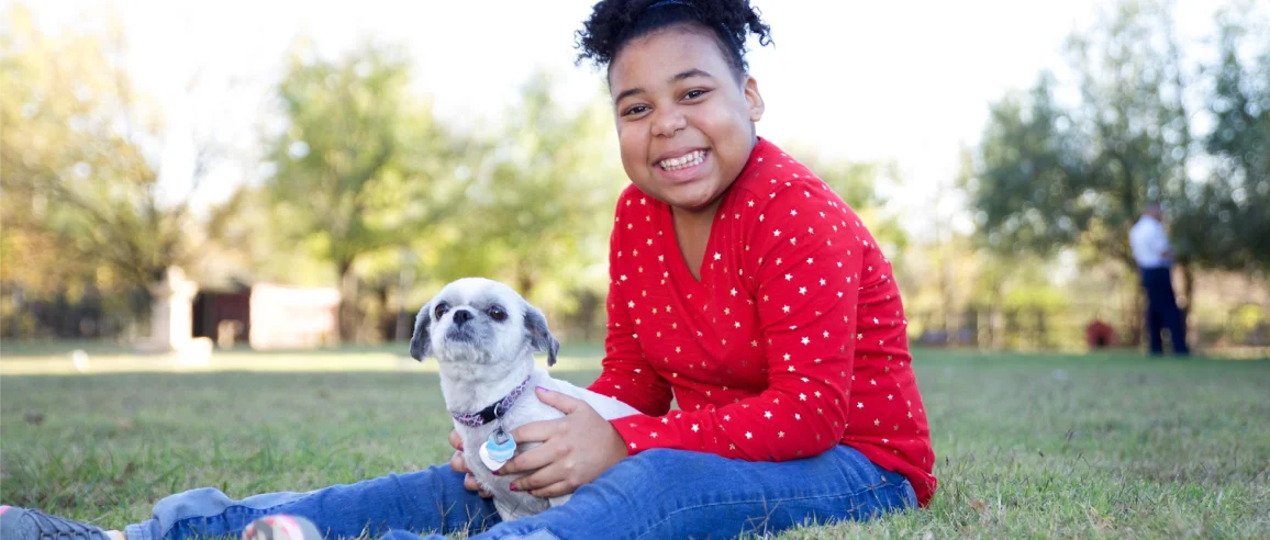 A smiling child in a red sweater sits on grass, gently holding a small dog. They are in a sunny park with trees in the background, conveying joy and warmth.