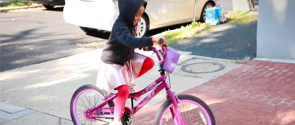 Un niño pequeño con una sudadera con capucha negra y falda rosa monta una bicicleta de color rosa brillante en una acera soleada. La escena es alegre, evocando una sensación de alegría y alegría.